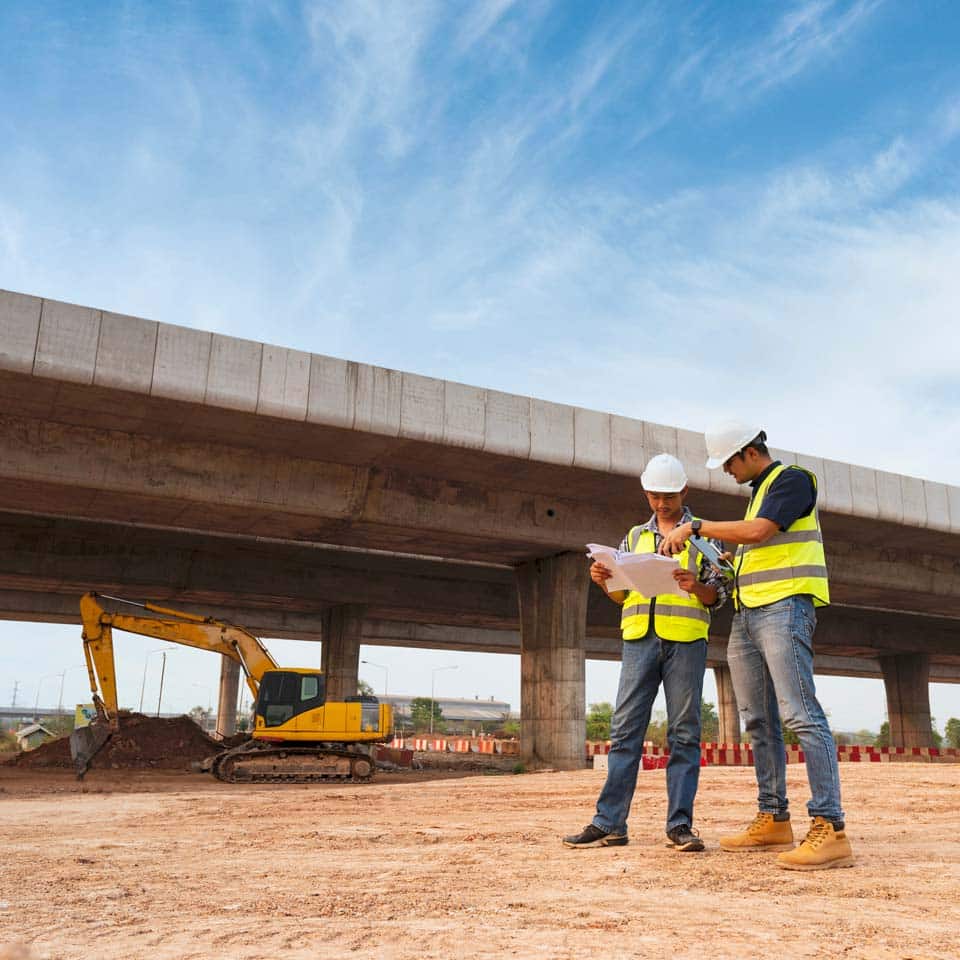 Two workers reviewing plans at a road construction project with a backhoe behind them.